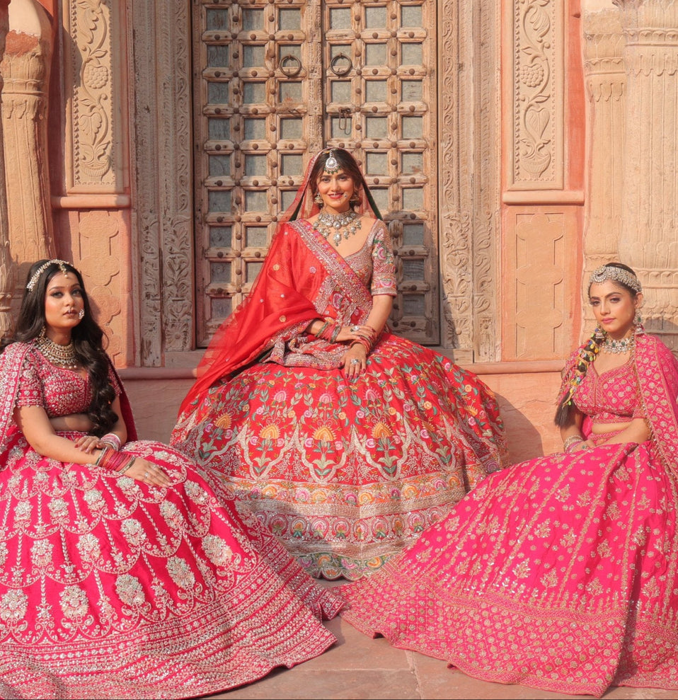 Five women in traditional pink and red lehengas sitting in front of a pink stone building.
